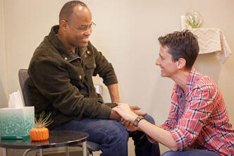 A Black man sits in a chair wearing jeans and a black jacket.  He is smiling.  Sloth Around acupuncturist Lynn kneels on the ground to take his pulse on his right hand.  She is wearing a red flannel button up shirt and smiling. 