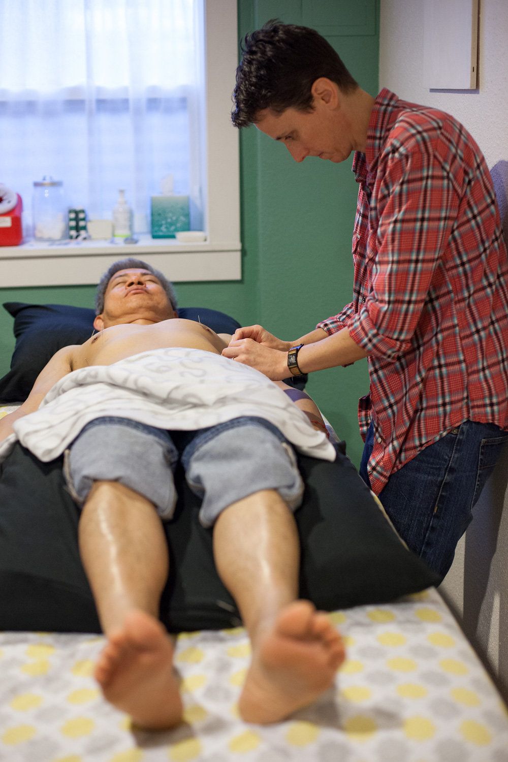 An Asian man in his fifties lays face up on a massage table.  He is not wearing a shirt and his jeans are rolled up to his knees.  He has a black pillow underneath his knees and head.  Acupuncturist Lynn is inserting an acupuncture needle into his left arm.  His eyes are closed and he appears calm.  