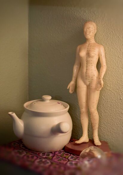 An artistic picture of three items associated with Traditional Chinese Medicine: a white teapot for cooking loose herbs, a plastic sculpture of a naked woman with meridian lines drawn on her body, and clear glass cups arranged on a table top with a brightly colored fabric underneath.