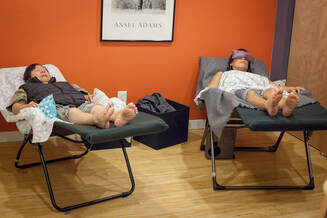 Two women recline in zero gravity chairs.  Both women have their pant legs rolled up to their knees and have acupuncture needles in their arms and legs.