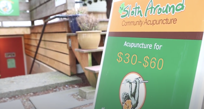 An orange and green A-Frame sign reads Sloth Around Community Acupuncture.  Off to the side, the entrance to the clinic is visible.  The clinic entrance has concrete steps and a black handrail on the right hand side.